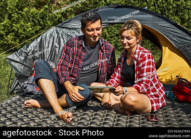 Couple planning next trip while sitting with map by tent. People relaxing at camping during summer vacation. Actively spending vacations outdoors close to...