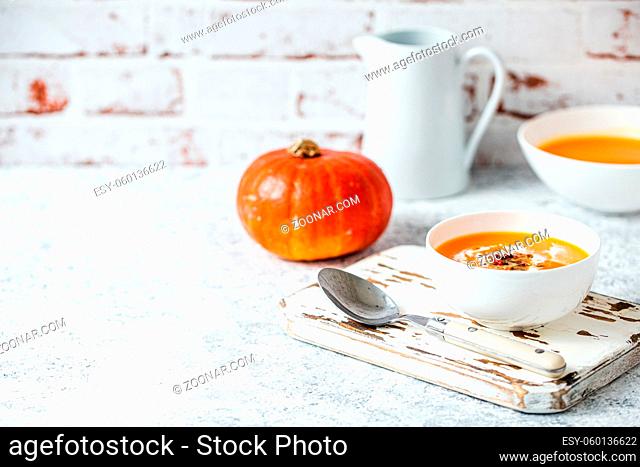 Homemade pumpkin cream soup served in white ceramic bowl on white table with spoon decorated with whole pumpkin, angle view, selective focus