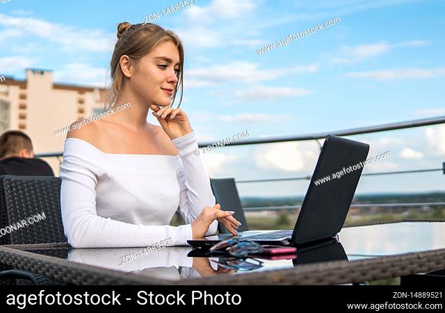 Beautiful young woman in white t-shirt is working on laptop and smiling while sitting outdoors in cafe. Young female using laptop for work