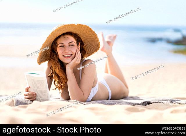 Smiling beautiful woman with book day dreaming while lying on sand during sunny day