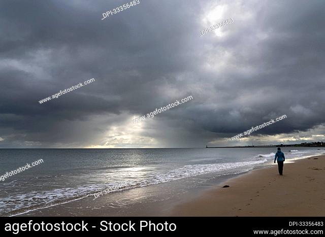 A woman walks on the sand beach at the water's edge under a dark cloudy sky; Whitburn, Tyne and Wear, England