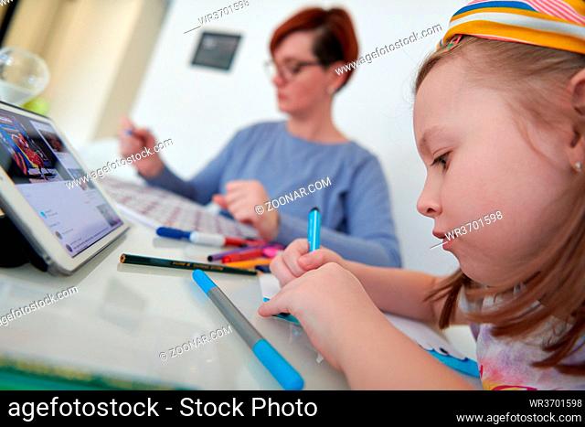 Mother and little daughter playing together drawing creative artwork during coronavirus quarantine measuring time on sandglass and listening music on tablet