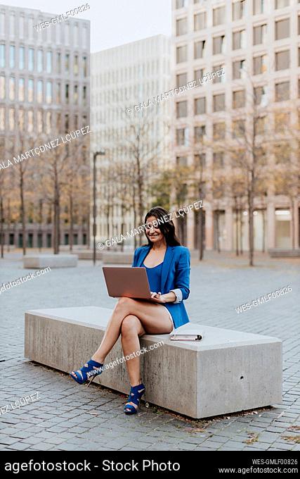 Happy businesswoman using laptop while sitting on bench in city