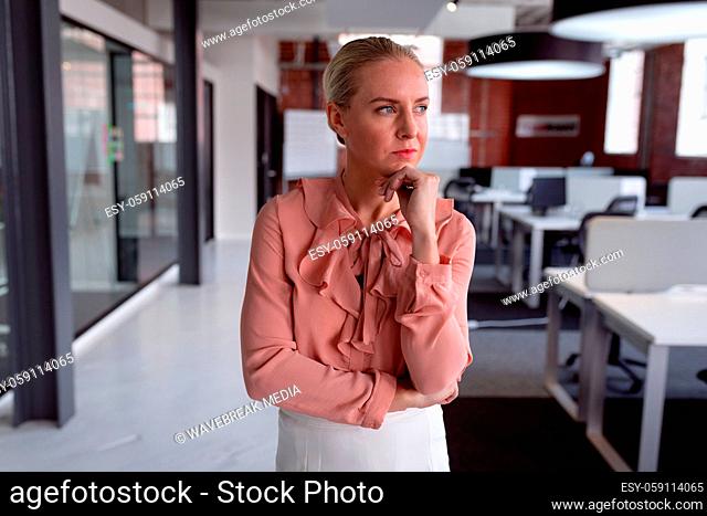 Thoughtful caucasian businesswoman standing in office with chin on hand, looking away