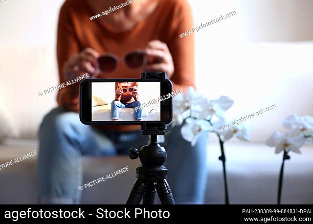08 March 2023, Bavaria, Kaufbeuren: A woman sits on a sofa with sunglasses in her hands while filming herself with her iPhone (posed scene)