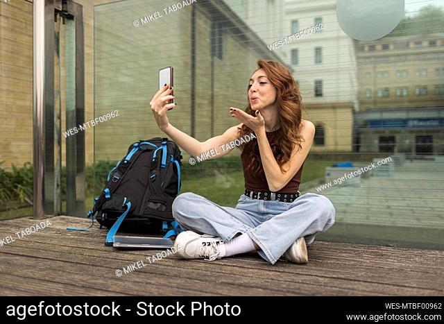 Woman blowing a kiss to video call through mobile phone in front of glass wall