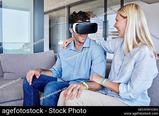 Cheerful woman with arm around looking at man wearing virtual reality simulator in living room