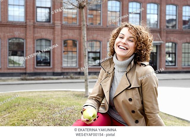 Portrait of happy woman with curly hair sitting on a meadow eating an apple