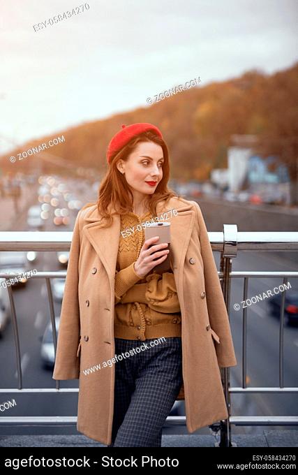 Tender young woman warming up with cup of coffee on the street female fashion standing on a pedestrian bridge. Portrait of stylish young woman wearing autumn...