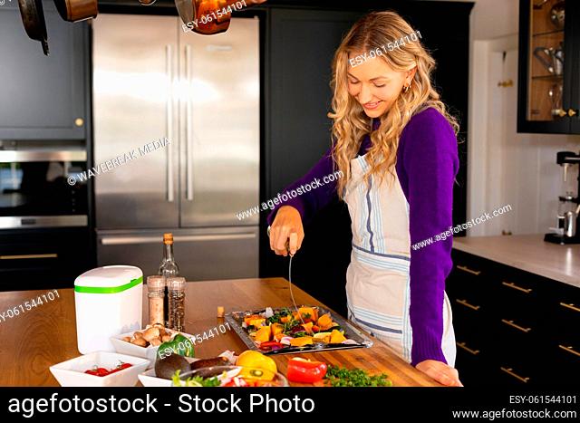 Smiling caucasian woman standing in kitchen in apron seasoning chopped vegetables