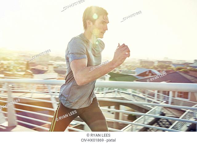 Determined male runner running on sunny urban footbridge at sunrise