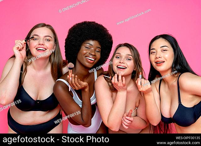 Cheerful multi-ethnic female models in lingerie holding beauty products against pink background