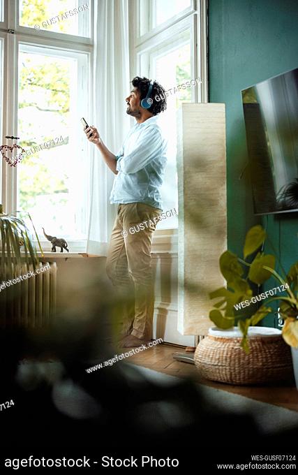 Man with smart phone listening music through wireless headphones by window at home