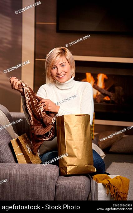 Happy young woman siiting on sofa at home taking out clothes of shopping bags. smiling