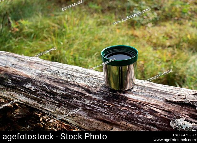 Thermos green plastic stainless steel mug put on a log on sunny day surounded by green grass in a forest