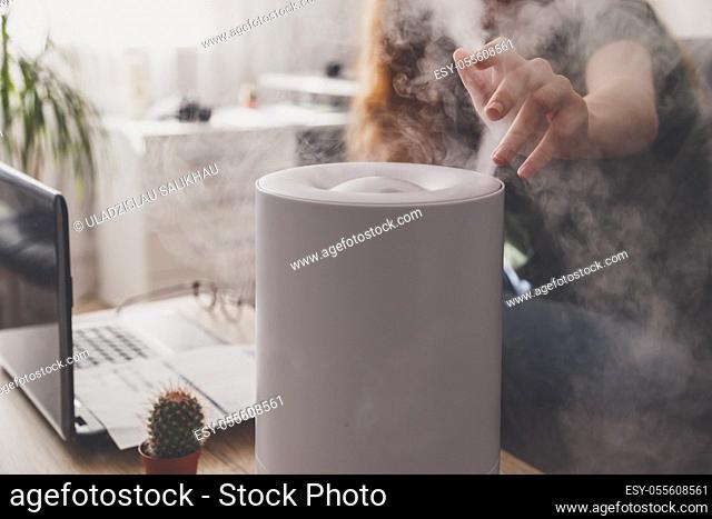 Woman freelancer uses a household humidifier in the workplace to maintain relative humidity and microclimate in the workplace of the home office with a laptop...