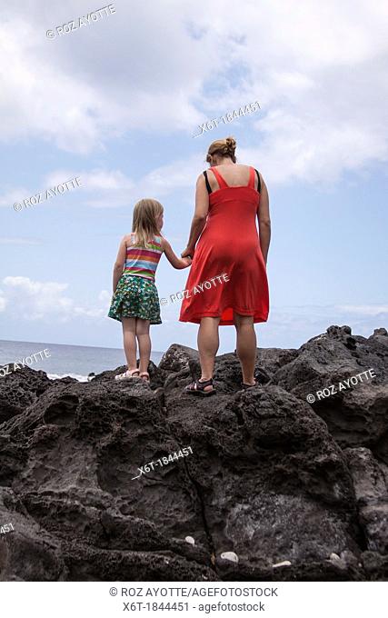 back of mother wearing a red dress, holding hands with a young girl in a striped shirt and a flower printed skirt  both are standing on giant lava rocks looking...