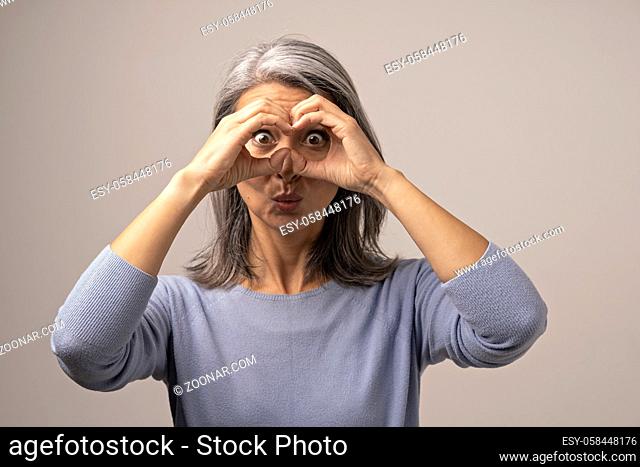 Mongolian Mature Woman Holds Hands As if Looking Through Binoculars. The Woman Has Gray Hair. Her Face Looks Funny. She Opened Her Eyes Wide with Surprise