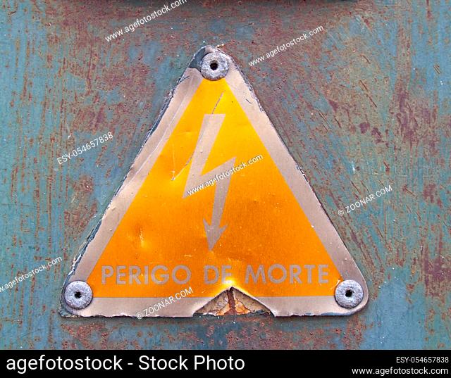 an old metal triangular yellow Portuguese electricity safety sign with a lightning bold symbol reading perigo de morte - translation danger of death on a rusty...