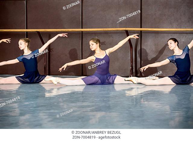 Young ballerinas on the lesson of classical choreography sitting in the splits gracefully raising his hands up near barre