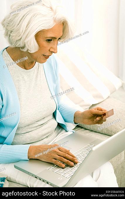 gray haired senior italian woman holds credit or debit card and use laptop to do online payments, or banking or shopping sitting on a white couch or sofa