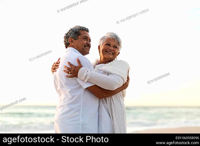 Smiling senior newlywed multiracial couple embracing while looking away at beach during sunset