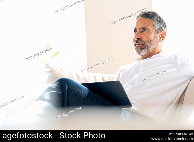 Thoughtful businessman sitting with digital tablet on sofa at home