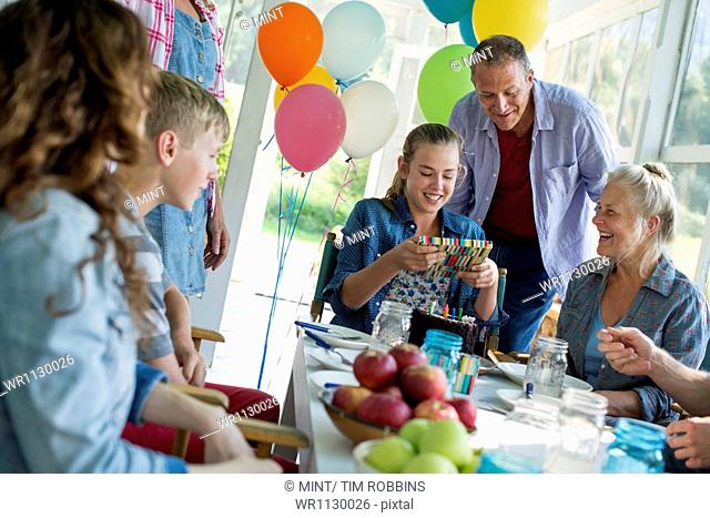 A birthday party in a farmhouse kitchen. A group of adults and children gathered around a chocolate cake