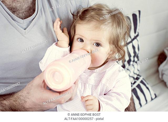 Baby girl sitting on father's lap, drinking from bottle, cropped