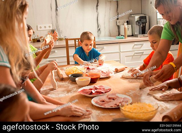 Two women and a group of children are sitting at a table in the kitchen. Women show children how to cook pizza