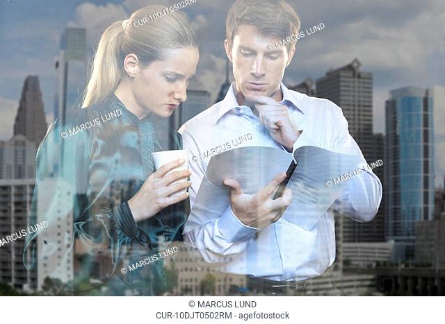 Young business man and woman looking out of window with reflection of city skyline, discussing a document