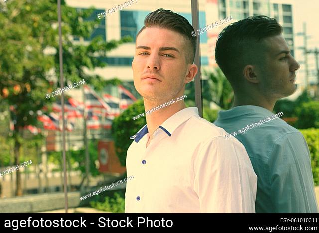 Young handsome man leaning against glass window of modern building in Bangkok Thailand
