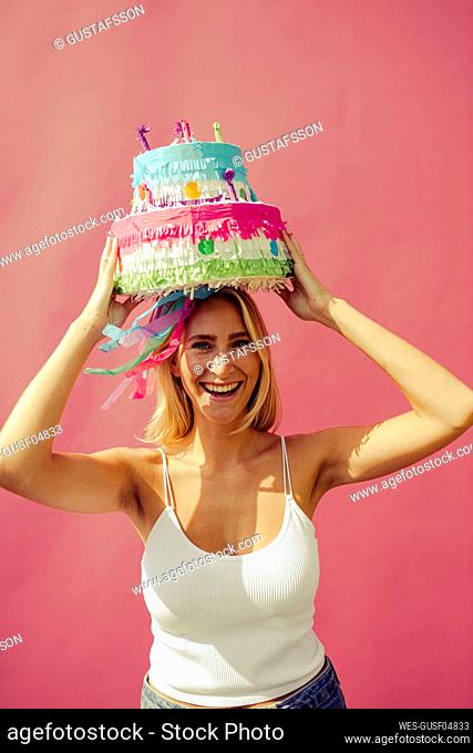 Cheerful woman carrying birthday cake on head against pink wall