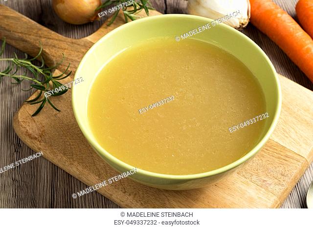 Chicken bone broth in a green soup bowl, with carrots, onions, and rosemary in the background