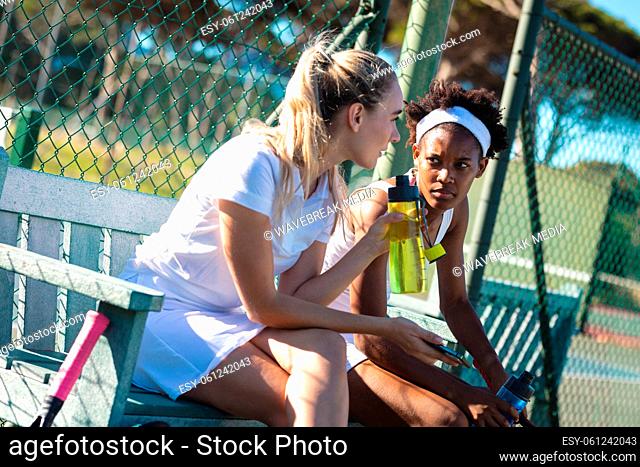 Young biracial female tennis players talking while sitting at bench in court during game break