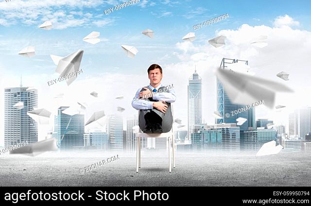 scared businessman sitting on a chair with his hands clasped his knees, paper plane fly around