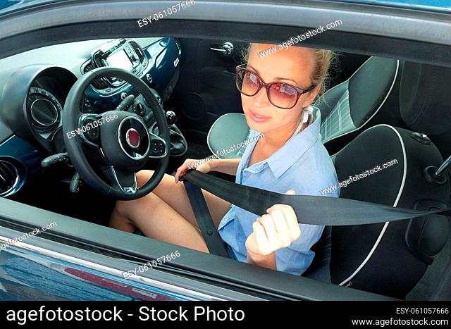 Safety first. Beautiful blonde casual caucasian woman wearing sunnglasses, blue shirt and jeans shorts, fastening car seat belt