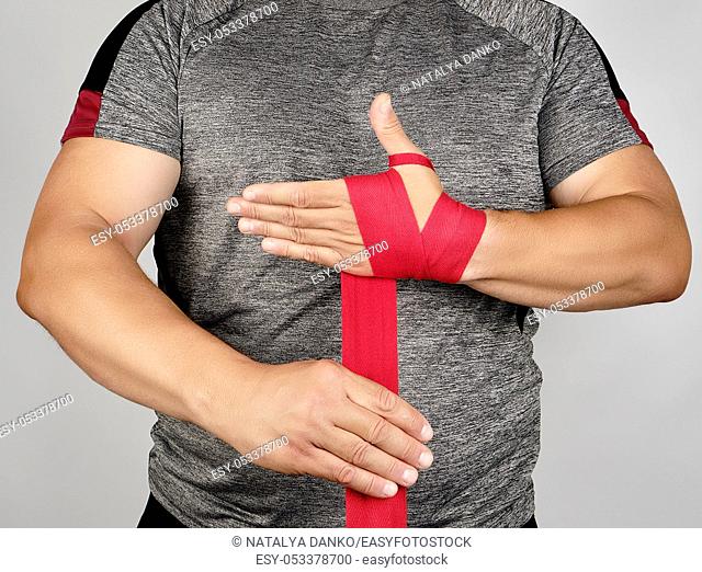 athlete stand in gray clothes and wrap his hands in red textile elastic bandage before training, gray background