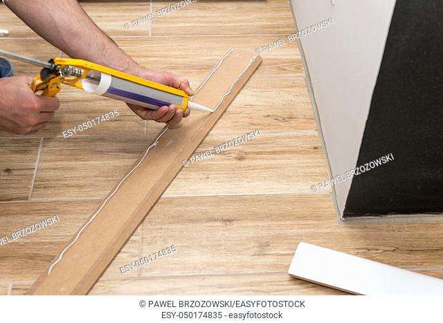 Mans hands installing white skirting board with caulking gun silicone from cartridge