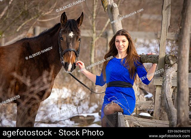 A beautiful girl is sitting on a log against the background of wooden ruins, a horse is standing nearby