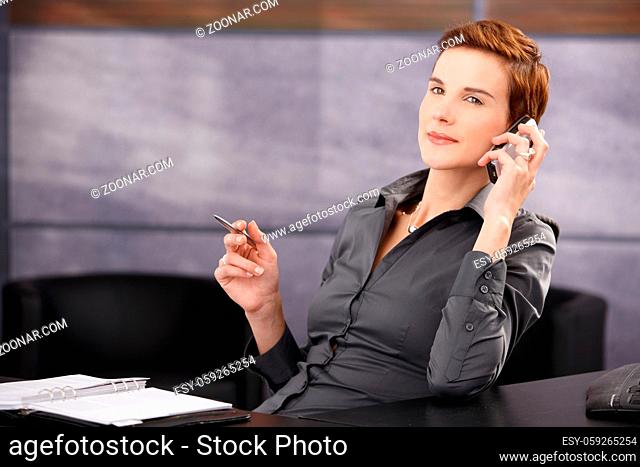 Confident businesswoman sitting at desk with pen handheld, concentrating on mobile phone call, smiling