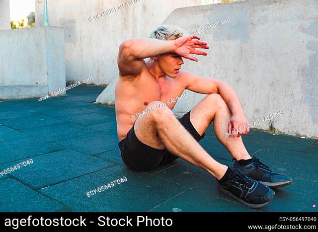 Young caucasian tired athlete man wipes sweat from forehead sitting on work out area outdoors