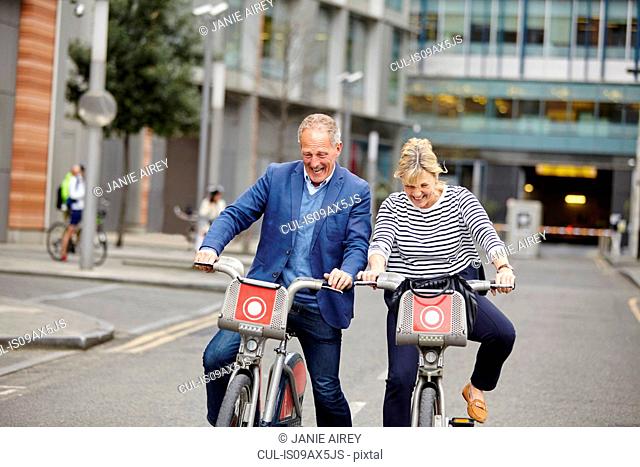 Mature dating couple laughing whilst cycling on hire bicycles, London, UK