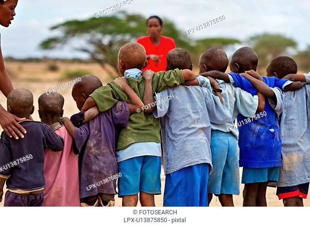 Masai school out on the plains: as seen from behind, a group of children, mostly dressed in blue sweaters, and blue form a tight line, with arms interlocked