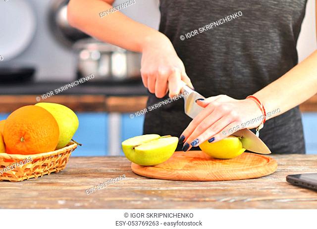 A young woman in a black t-shirt cuts green apple on a light wooden board in a kitchen. Fruit in bowl is standing near. Horizontal photo