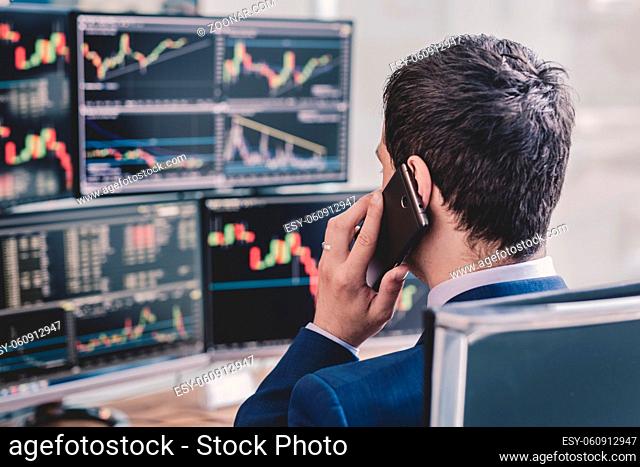 Over the shoulder view of and stock broker trading online while accepting orders by phone. Multiple computer screens ful of charts and data analyses in...