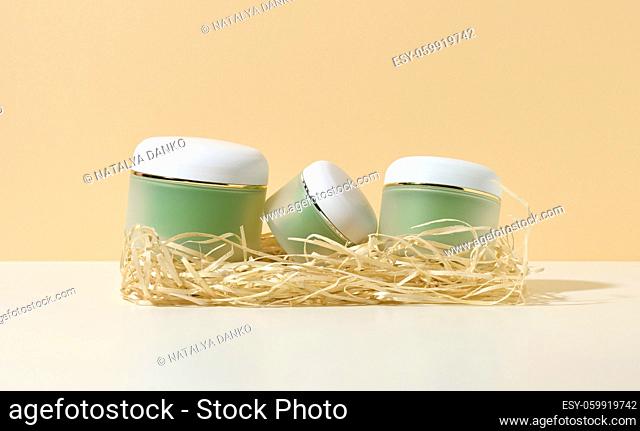 empty cosmetic green jars with white lids lie in wood shavings on a white table, beige background. Blank for branding products cream, gel, serum