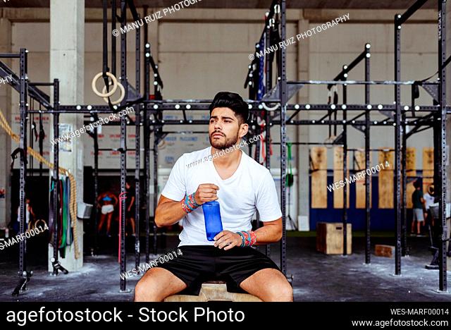 Young man holding water bottle while sitting on box at gym