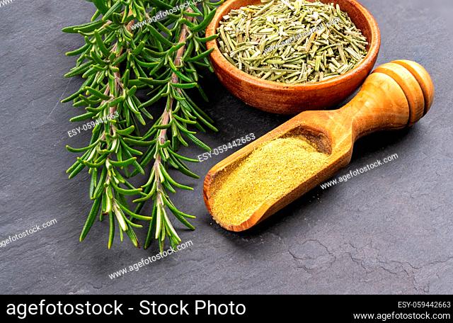 Fresh rosemary twigs, a spice shovel with ground rosemary and a wooden bowl with dried rosemary on a black slate background