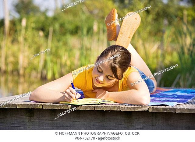 Teenaged girl lying on footbridge and writing in her diary with a slow smile